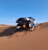 Geländefahrzeug hat sich auf einer Sanddüne in der Wüste festgefahren, mehrere Personen versuchen, das Fahrzeug freizuschaufeln