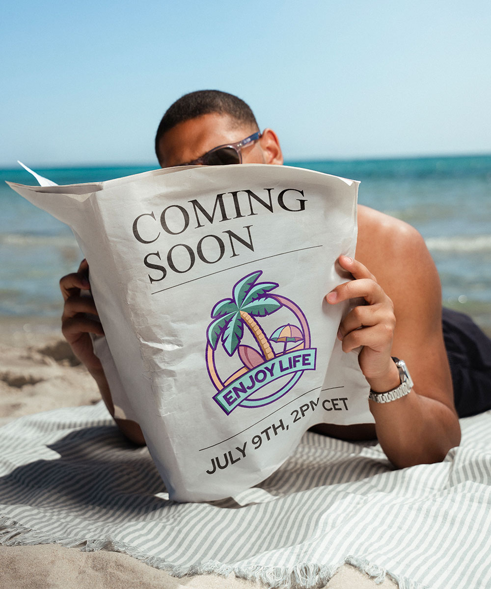 Freedom GMT Enjoy Life Holiday Limited Edition von Norqain am Handgelenk eines Mannes, der am Strand liegt und eine Zeitung liest mit der Aufschrift Coming Soon
